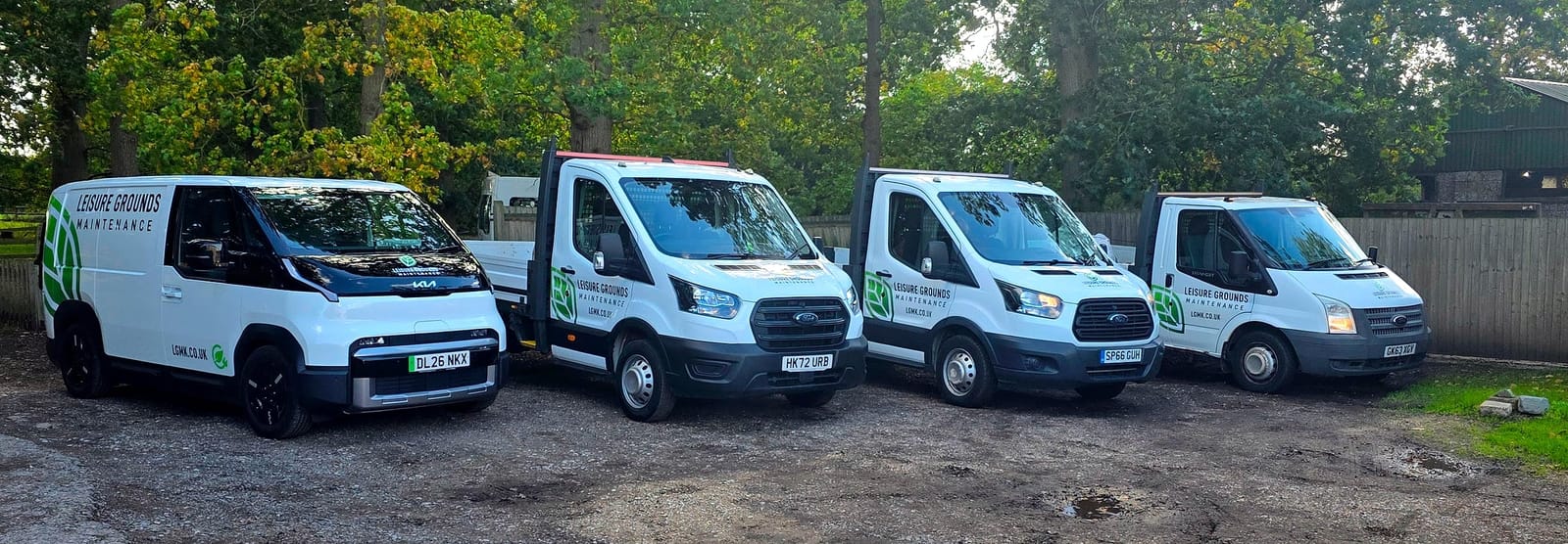 The LGM fleet parked in a row in front of trees, all in the green leaf livery with lgmk.co.uk on the side: the fully electric Kia van on the left and Ford tipper trucks beside it.